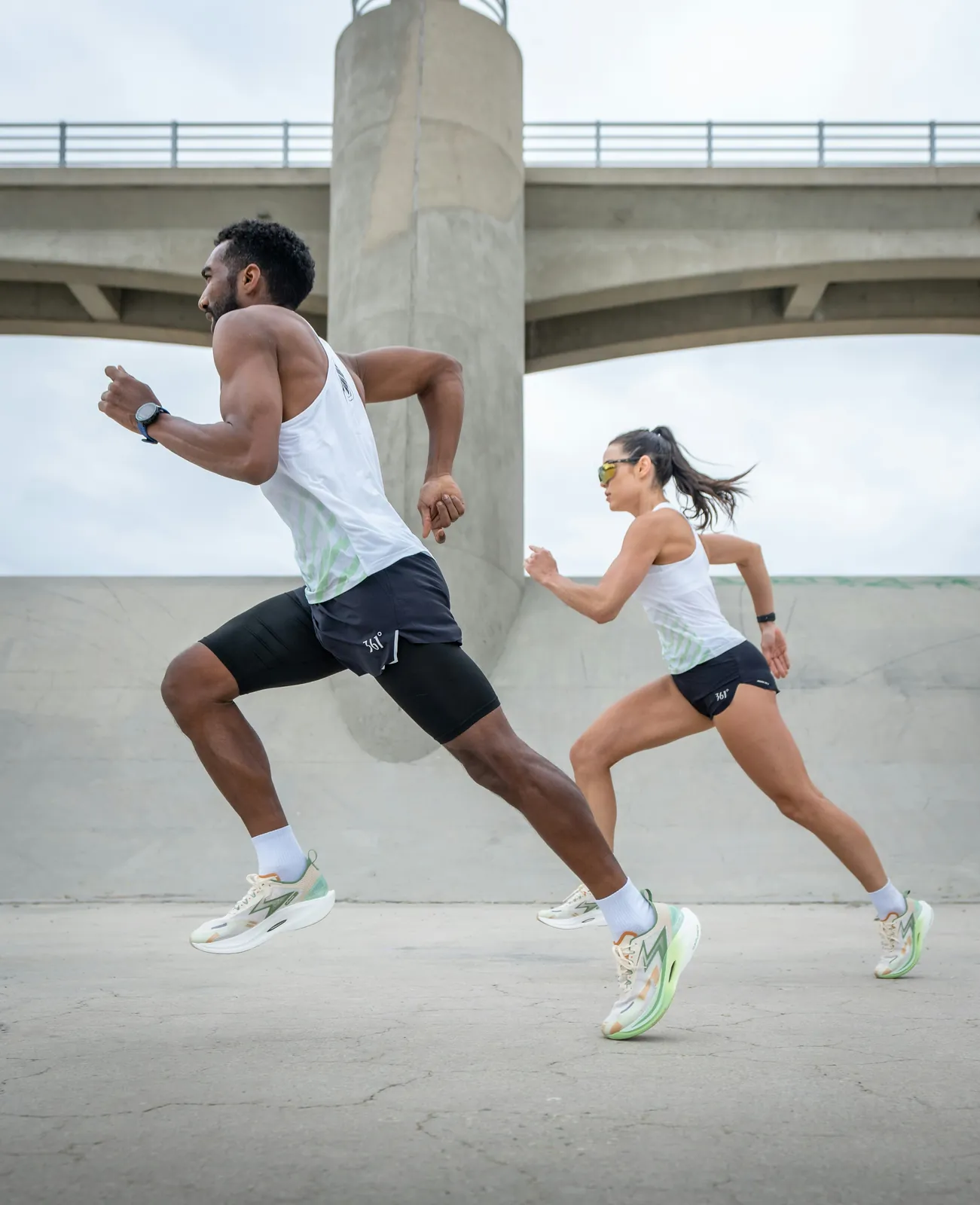 Two runners sprinting side by side on pavement beneath a concrete bridge on an overcast day